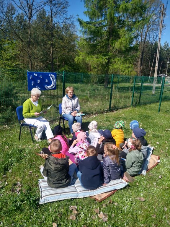 Dzieci siedzące na kocyku w ogrodzie słuchają książek czytanych przez bibliotekarkę i wolontariuszkę.