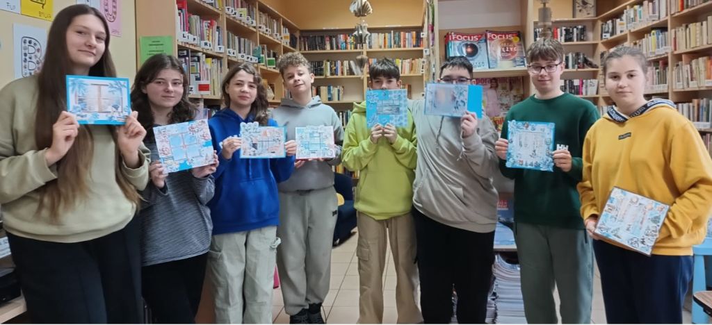 Uczestnicy warsztat&oacute;wstoją na tle bibliotecznych regał&oacute;w i demonstrują swoje prace.