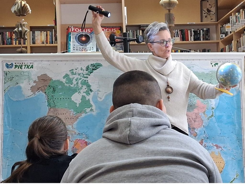 Bibliotekarka, stojąc przed uczniami na tle mapy, trzymając w jednej ręce globus i w drugiej latarkę, demonstruje oświetlanie Ziemi przez słońce.
