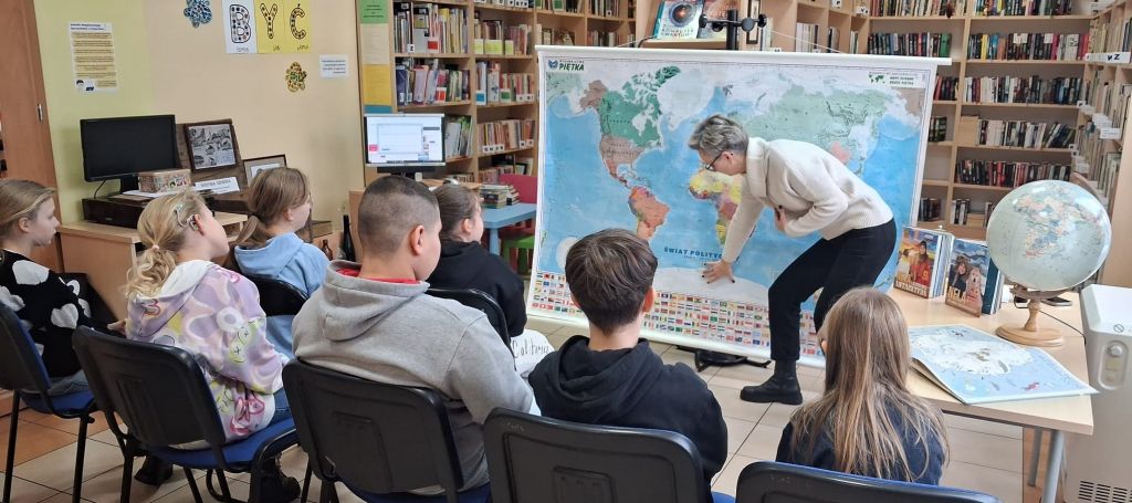 Grupa dzieci, siedzących na krzesłach przygląda się bibliotekarce, kt&oacute;ra pochylona pokazuje na mapie położenie Antarktydy.