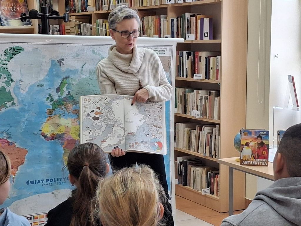 Bibliotekarka, trzymając w rękach otwartą książkę: Mapy. Obrazkowa podr&oacute;ż po lądach, morzach i kulturach świata, wskazuje palcem mapkę Arktyki.