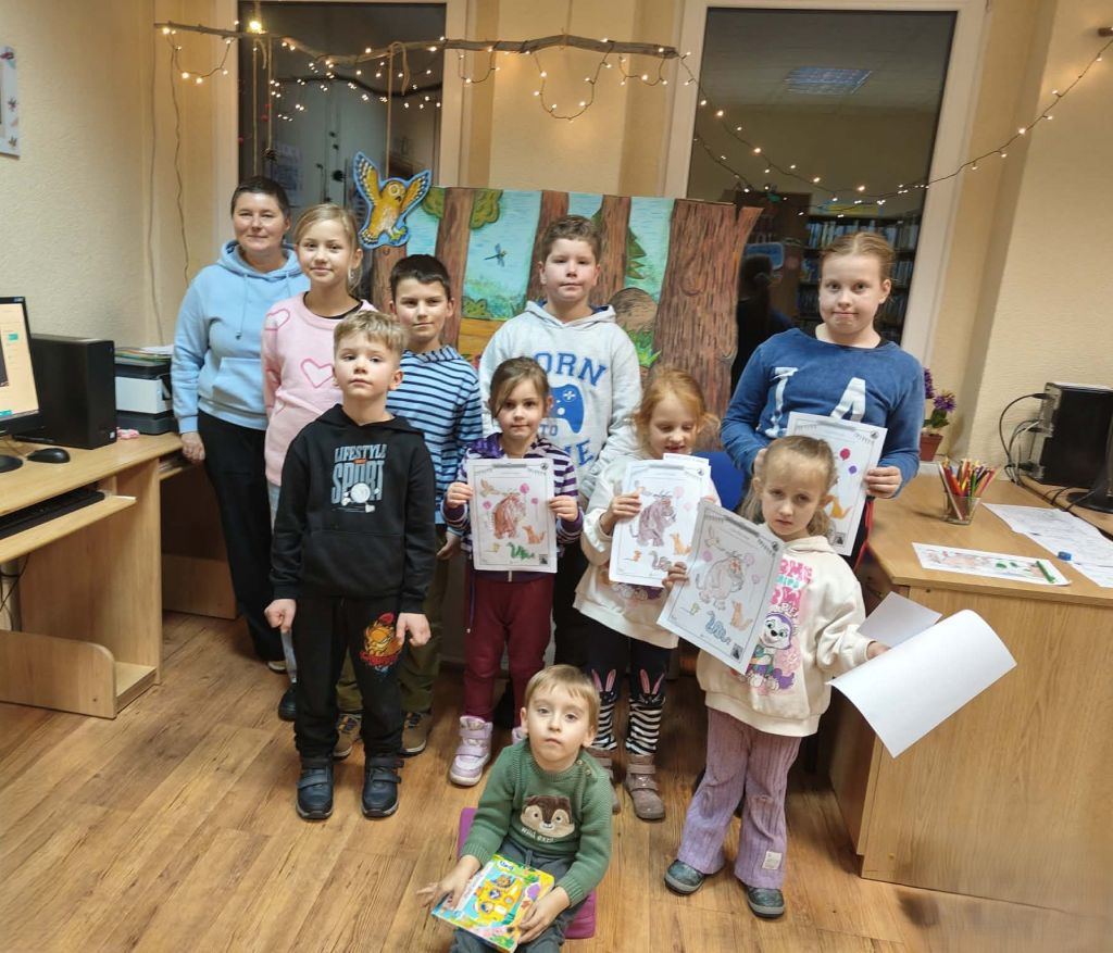 Stojąca grupa dzieci wraz z panią. Dzieci prezentują praca plastyczne, kolorowanki