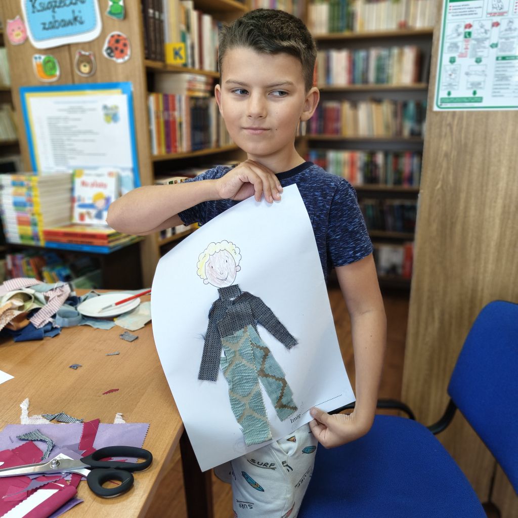Chłopiec trzyma pracę plastyczną pokazującą postać chłopca do której doklejone są kawałki materiału w formie ubrania. Chłopiec stoi na tle regałów bibliotecznych.