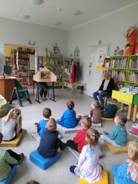 Bibliotekarz czyta bajkę w formie teatrzyku kamishibai, dzieci siedzą na poduchach