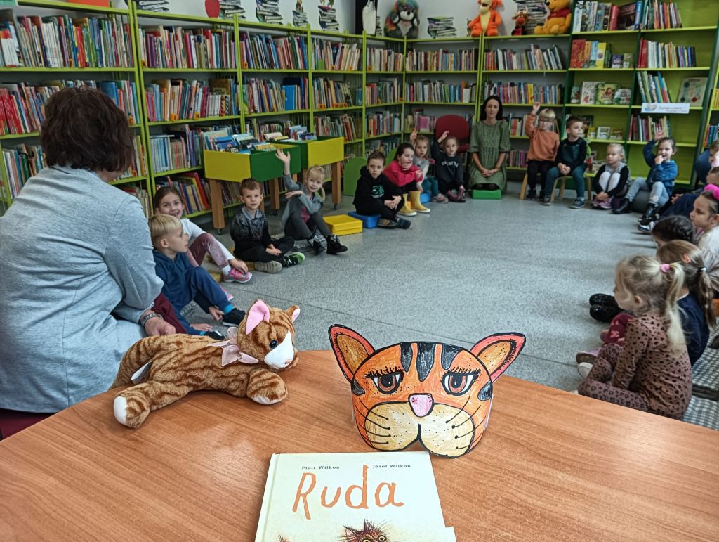 Dzieci siedzą w kole, bibliotekarz rozmawia z dziećmi, na stole leży książka Ruda