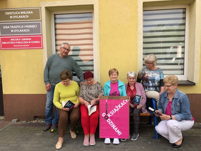 Członkowie Dyskusyjnego Klubu Książki siedzą przed budynkiem biblioteki. Jedna osoba trzyma plakat promocyjny DKK