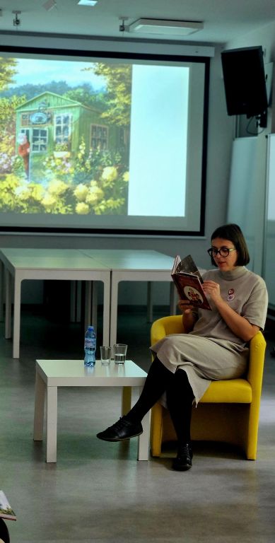 Agnieszka Blazy siedzi na fotelu i czyta dzieciom książkę Wnuczka antykwariusza