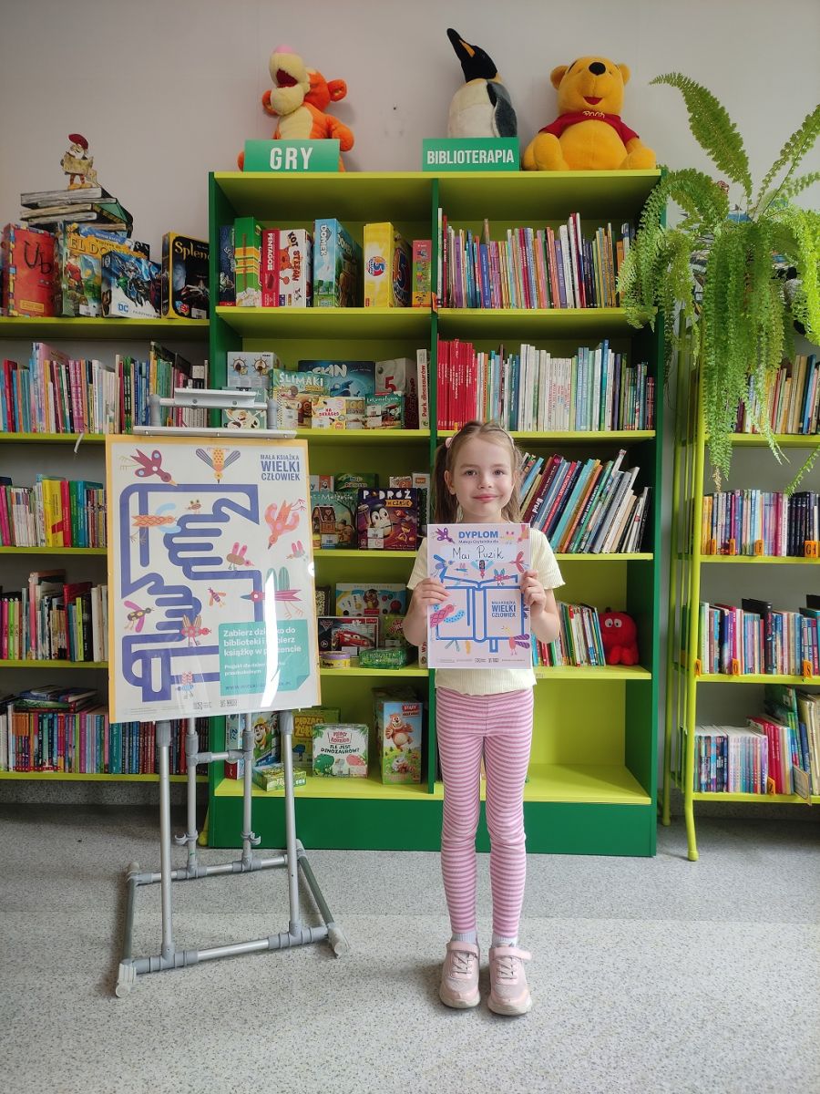 Dzieczynka trzyma zdobyty dyplom i stoi na tle regał&oacute;w w bibliotece.