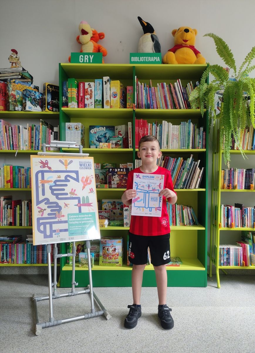 Chłopczyk trzyma zdobyty dyplom i stoi na tle regał&oacute;w z książkami w bibliotece.