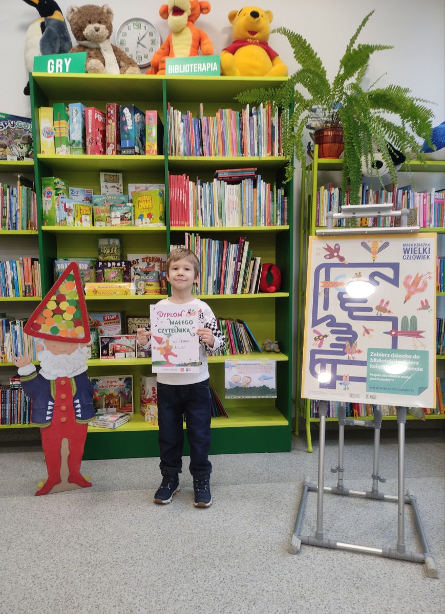 Chłopczyk pozuje do zdjęcia trzymając zdobyty dyplom z projektu "Mała książka- wielki człowiek" na tle regał&oacute;w z książkami.