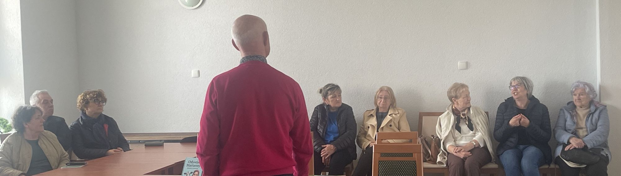 W rzędzie siedzą uczestnicy spotkania, przed kt&oacute;rymi stoi pan Zbigniew Stańczak. Pisarz wraz z pozostałymi przysłuchuje się wypowiedzi jednej z uczestniczek.