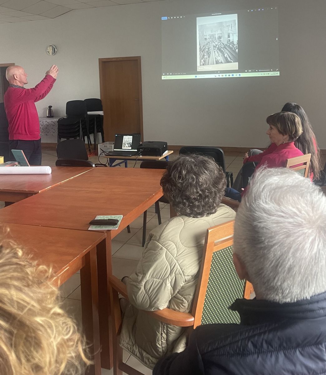 Na pierwszym planie kilkoro czytelnik&oacute;w, siedząc przy stole spoziera na wyświetlaną na ścianie prezentację. W głębi widać autora, wskazującego ręką slajd. 