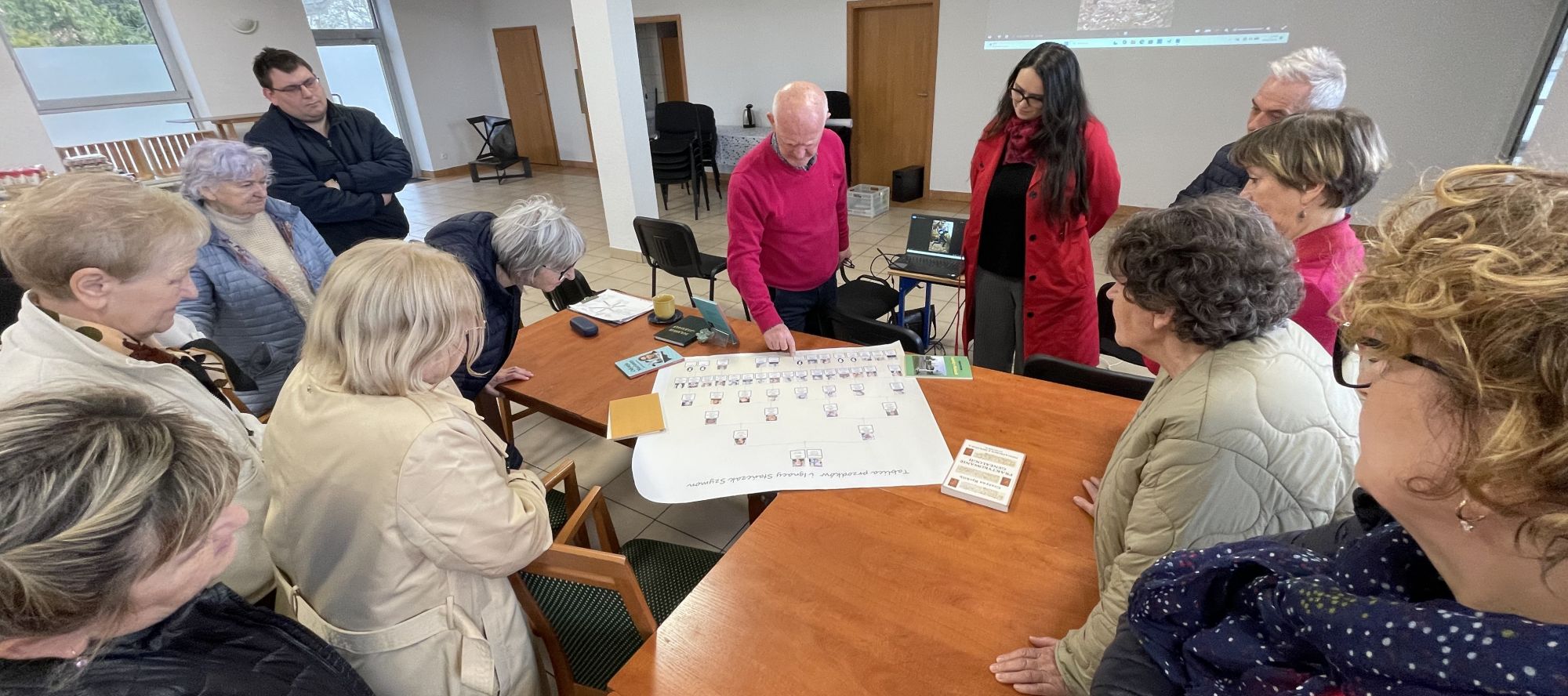 Cała grupa uczestnik&oacute;w stoi wokoło stołu, na blacie kt&oacute;rego rozpostarta jest tablica drzewa genealogicznego pisarza. Pan Stańczak wskazuje palcem miejsce tytułowej Marianny.