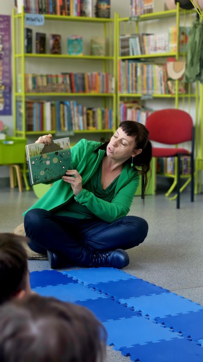 Ubrana na zielono bibliotekarka siedzi na podłodze i czyta dzieciom książkę o życiu żab.
