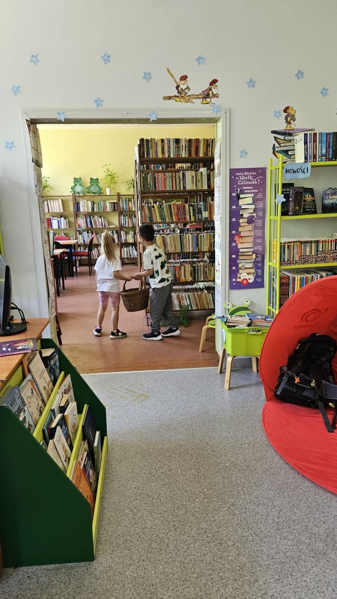 Chłopiec i dziewczynka idą razem przez bibliotekę trzymając wiklinowy koszyk na zajęciach poświęconych biwakowaniu. W tle regały z książkami