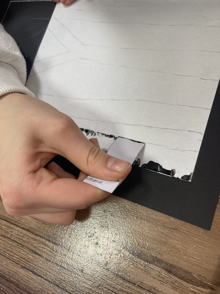 Zbliżenie na dłoń dziecka, kt&oacute;re z pomocą plastikowej karty skrobie na kartce czarny pień brzozy.