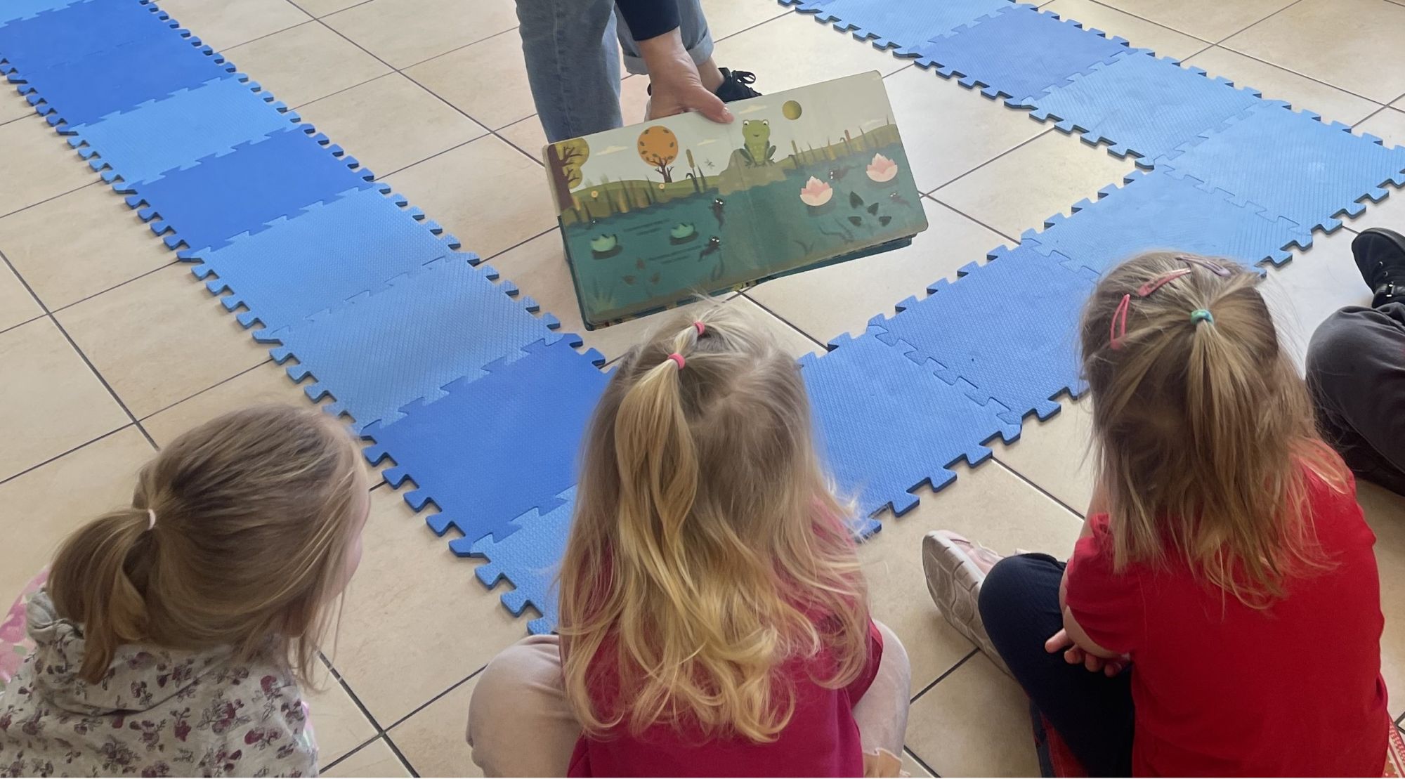 2-	Bibliotekarka, trzymając w dłoni otwartą książeczkę, prezentuje ją trzem dziewczynkom, siedzącym na podłodze.