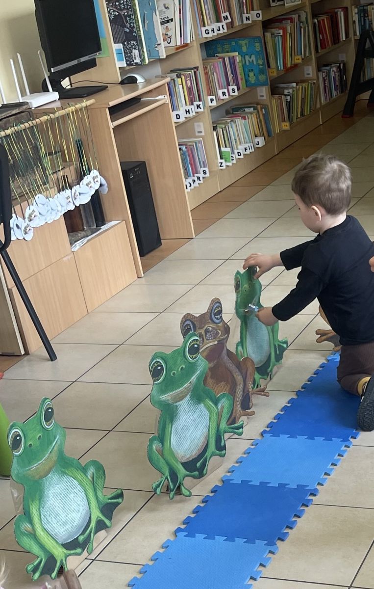 4-	Chłopczyk, klęcząc przed kartonową żabą, wkłada do jej pyszczka papierową muszkę.