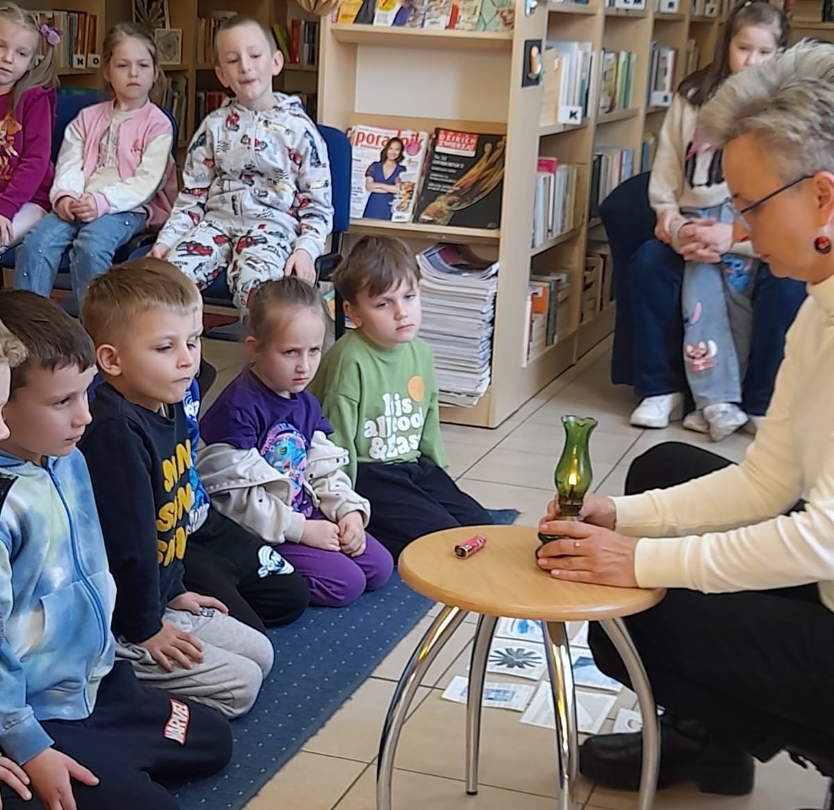 Grupie dzieci, siedzących na dywanie i krzesełkach, bibliotekarka prezentuje starą lampę naftową.