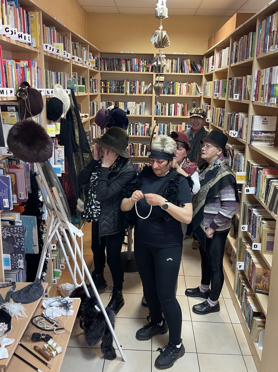 Grupa dorosłych, stojąc w bibliotece między regałami zakłada na siebie garderobę z stylu retro.