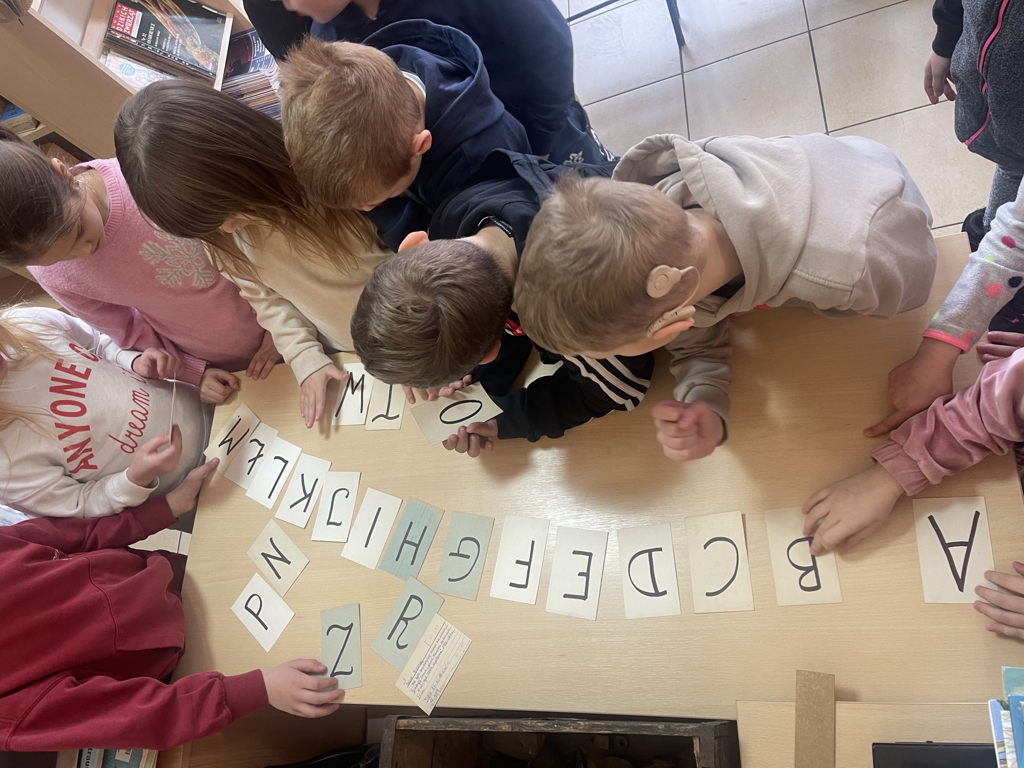 Grupa dzieci, stojąc wokoło stolika, na jego blacie układa polski alfabet.