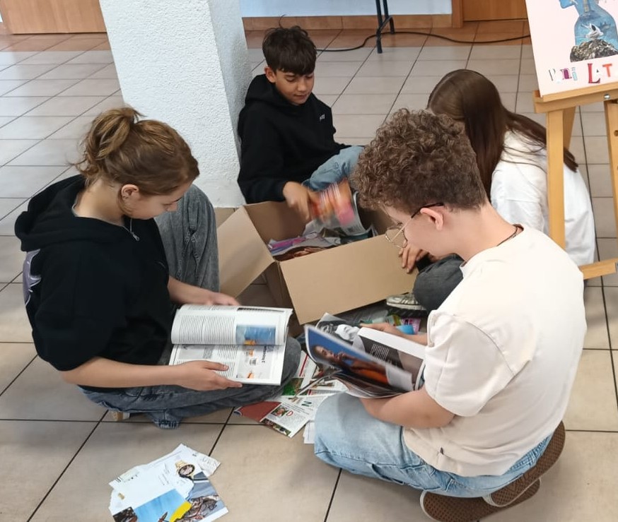 b-	Czw&oacute;rka uczni&oacute;w, siedząc na podłodze wokoło kartonu wertuje wyjęte z niego gazety.