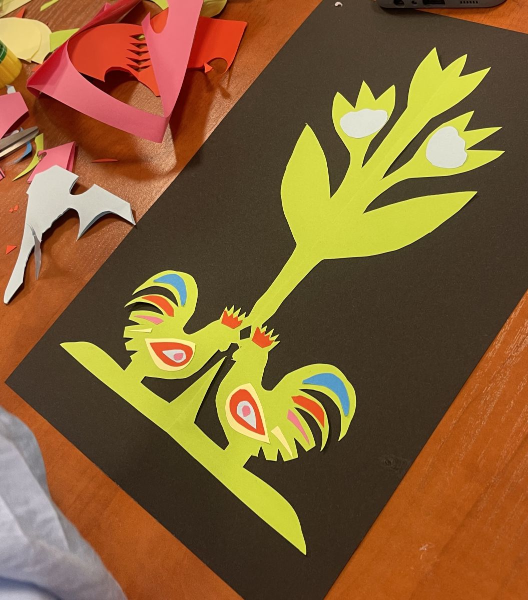 Zbliżenie na jedną z prac, leżącą na blacie stołu. Jest to jaskrawo ż&oacute;łta wycinanka  kwitnącej rośliny, pod kt&oacute;rą z obu stron są rozmieszczone symetrycznie dwa kogutki. Wycinanka zaprezentowana jest na brązowej kartce papieru.