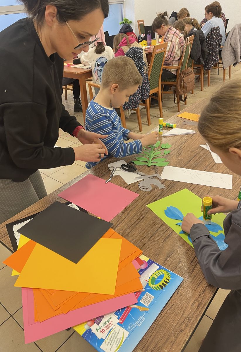 Na pierwszym planie przy stole stoi kobieta i dw&oacute;jka dzieci, kt&oacute;rzy przyklejają swoje wycinanki do kolorowych kartek. Za nimi w oddali st&oacute;ł gł&oacute;wny, przy kt&oacute;rym pracuje pozostała grupa uczestnik&oacute;w.