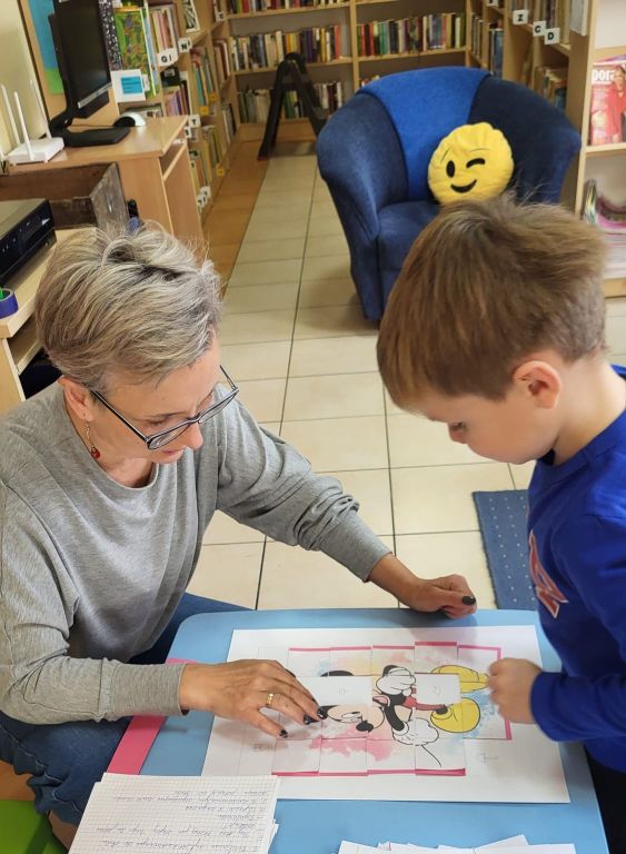 W sali bibliotecznej, na tle regał&oacute;w, przy małym stoliku bibliotekarka wraz chłopczykiem pochyleni są na puzzlami.