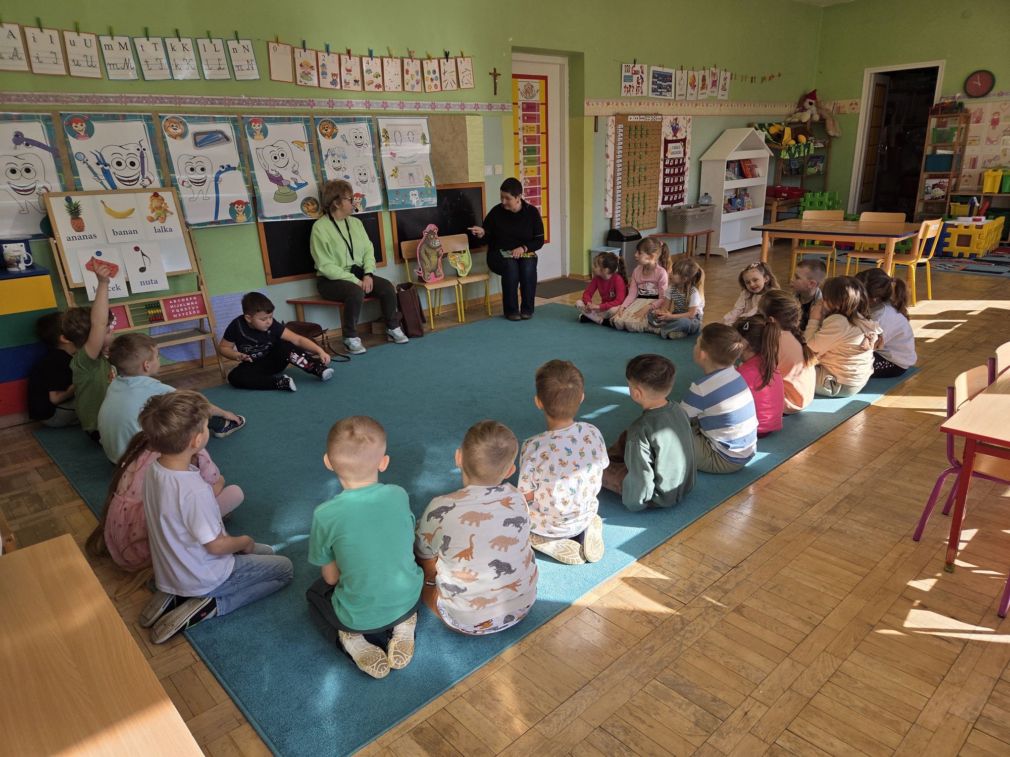  Dzieci siedzą w kole na dywanie i patrzą jak pani pokazuje im postaci z bajki.