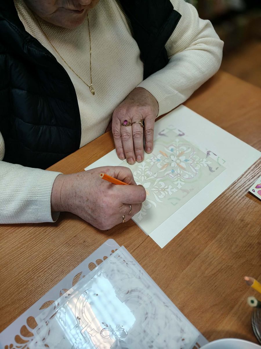 Na stole leży kartka papieru z rozpoczętą ilustracją ze wzorem w stylu folkowym. Widać ręce osoby rysującej na papierze.