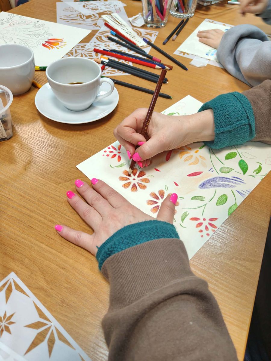 Na stole leży kartka papieru z rozpoczętą ilustracją ze wzorem w stylu folkowym. Widać ręce osoby rysującej, filiżankę z kawą i kredki. 