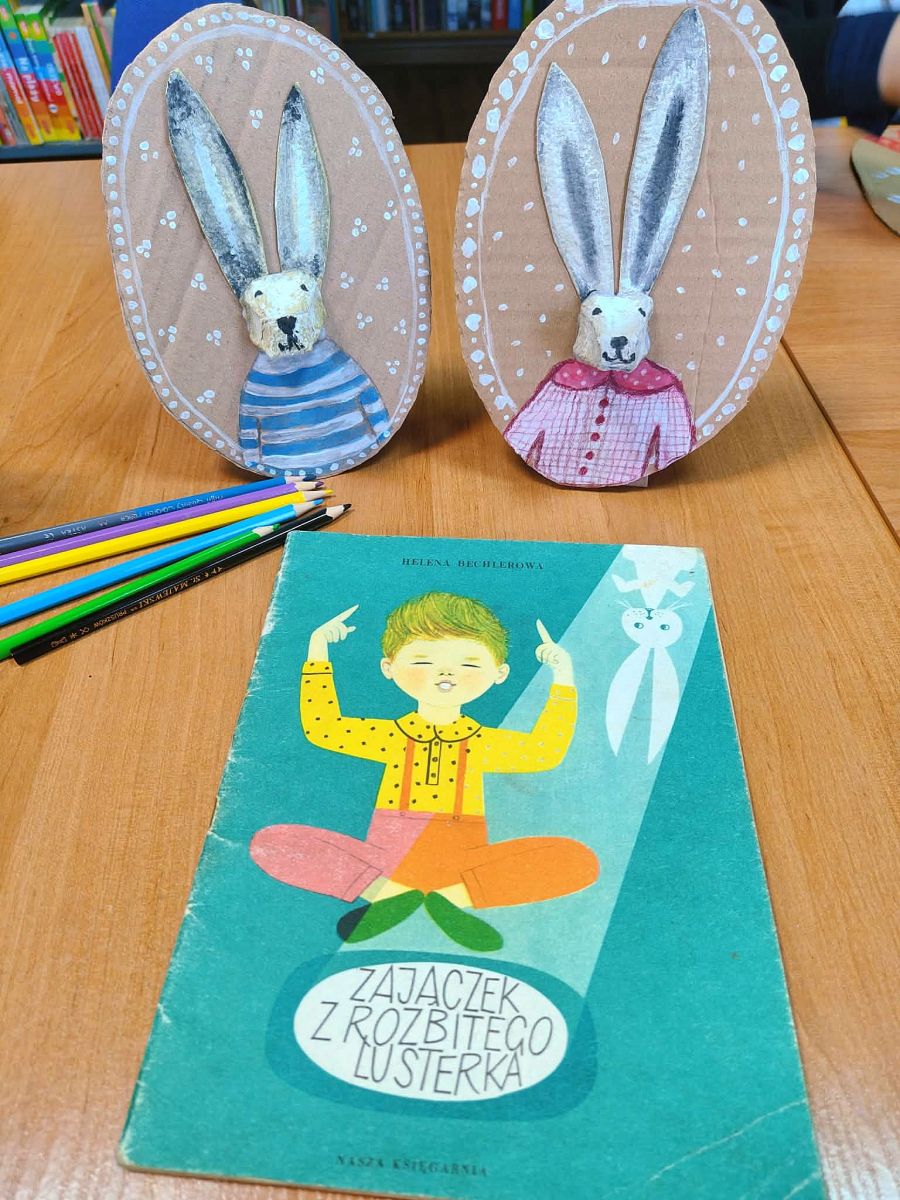 Na stole leży książka pt. 'Zajączek z rozbitego lusterka' a za nią dwa portrety zajęcy wykonane z tektury.