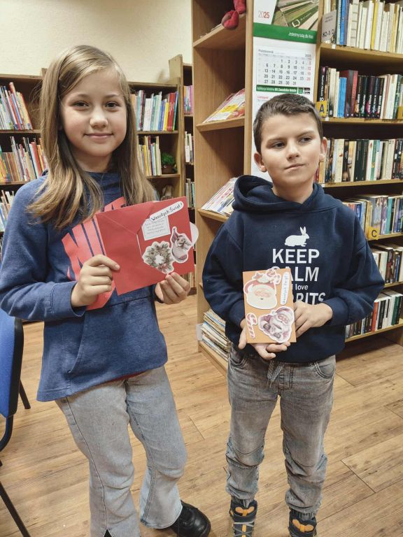 Na zdjęciu widać dwoje dzieci stojących w bibliotece. Oboje trzymają w rękach własnoręcznie wykonane kartki świąteczne ozdobione naklejkami i rysunkami. Po lewej stronie stoi dziewczynka w niebieskiej bluzy, z długimi, rozpuszczonymi włosami. Uśmiecha się lekko i trzyma czerwoną kartkę z zimowo-świątecznymi dekoracjami. Po prawej stronie stoi chłopiec w granatowej bluzie z napisem i kr&oacute;tkimi włosami. Trzyma pomarańczową kartkę udekorowaną m.in. wizerunkiem Mikołaja i świątecznymi elementami. W tle widać regały pełne książek oraz kalendarz na ścianie, co wskazuje, że dzieci znajdują się w czytelni lub bibliotece. Atmosfera jest spokojna i tw&oacute;rcza, jak podczas warsztat&oacute;w plastycznych.
