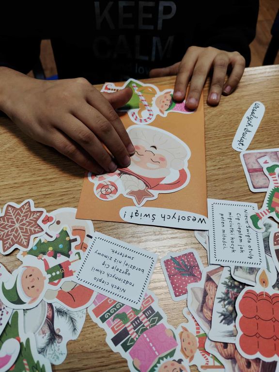 Na zdjęciu widać osobę, kt&oacute;ra własnoręcznie przygotowuje świąteczną kartkę. Na drewnianym stole leży pomarańczowa kartka ozdobiona naklejką uśmiechniętego Mikołaja. Ręce osoby delikatnie dociskają elementy dekoracyjne. Wok&oacute;ł porozrzucane są r&oacute;żne kolorowe naklejki o tematyce świątecznej: gwiazdki, prezenty, elf, choinki oraz małe karteczki z życzeniami po polsku, m.in. &bdquo;Wesołych Świąt!&rdquo;. Całość sprawia wrażenie miłej, kreatywnej pracy nad bożonarodzeniową kartką.