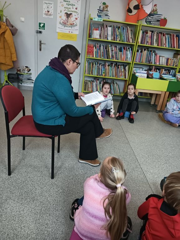 Bibliotekarz siedzi i czyta dzieciom opowieść, dzieci siedzą w półokręgu i słuchają