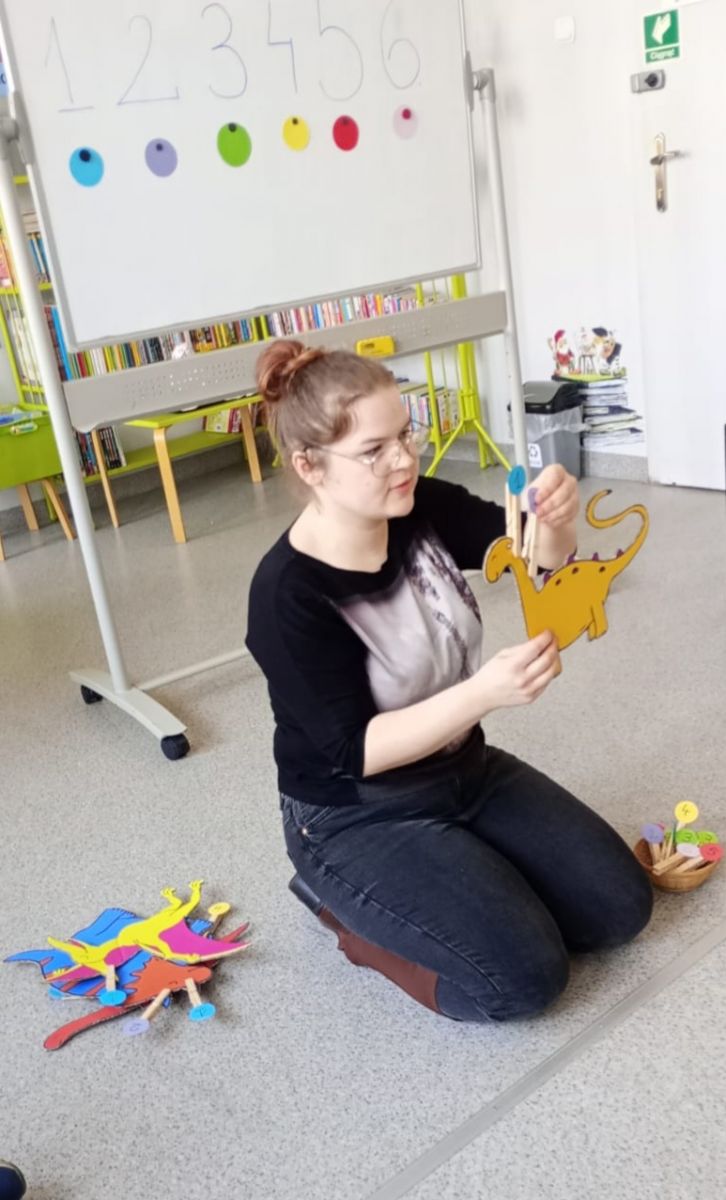 Prowadząca zajęcia liczy z dziećmi płytki grzbietowe dinozaur&oacute;w przyczepiając klamerki z cyferkami na każdym z nich. Nauka liczenia.