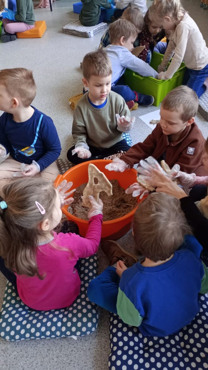 Grupa przedszkolak&oacute;w wykopuje z piski pełenj piachu kości dinozaura wykonane z masy solnej.