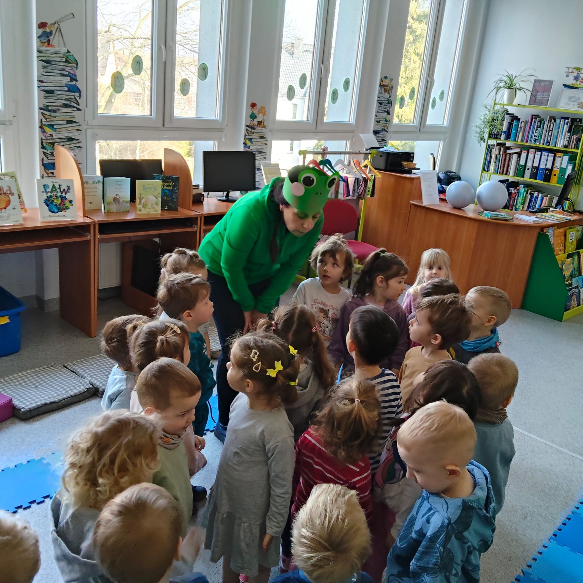 Przedszkolaki wraz z Panią zebrały się na środku sali w k&oacute;łeczko i bawią się w udawanie kijanek.