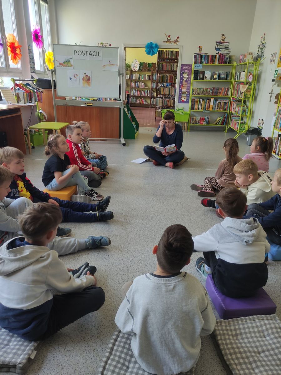 Pani bibliotekarka czyta fragment ksiązki zwr&oacute;cona do grupy dzieci.