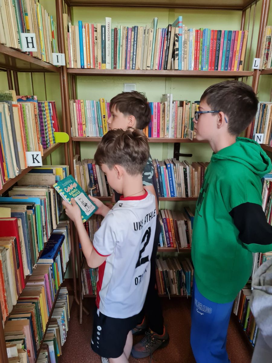 Trzech uczni&oacute;w rozglada sie po p&oacute;łce z książkami w poszukiwaniu ciekawych dla nich publikacji.