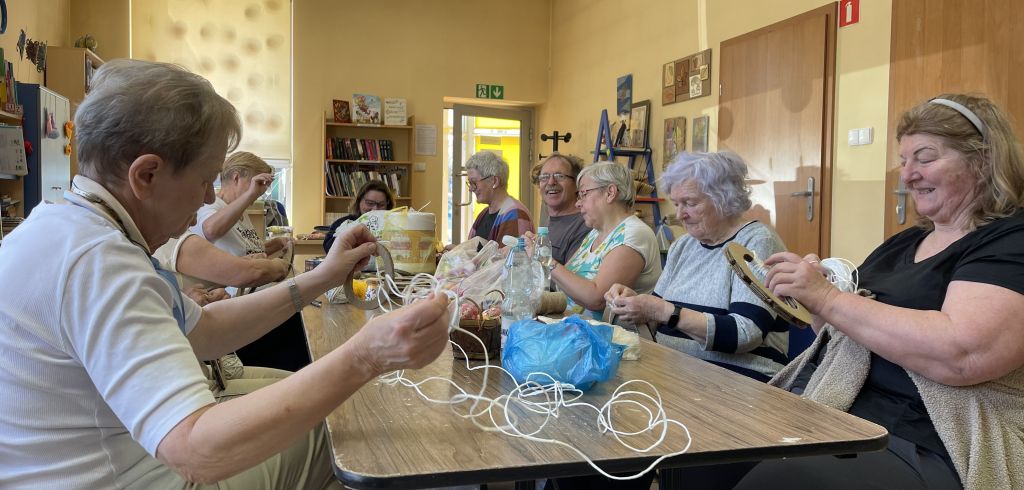 Grupa seniorów podczas warsztatów w bibliotece wykonuje ozdobę świąteczną ze sznurka, wełny i elementów dekoracyjnych.